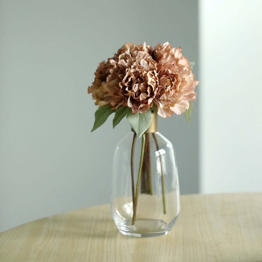 Pink artificial flowers in a clear glass vase on a wooden table.