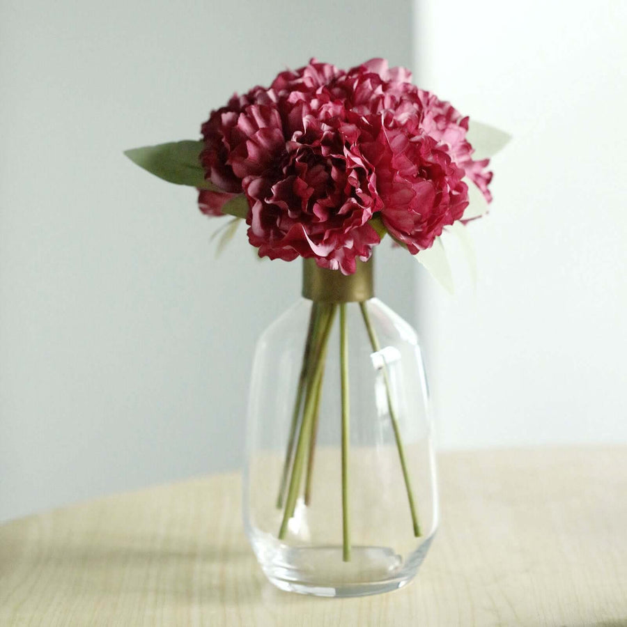 Artificial red peony bouquet in a clear vase on a wooden table.