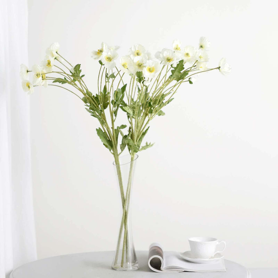 White flowers in a clear vase on a table with a cup and magazine.