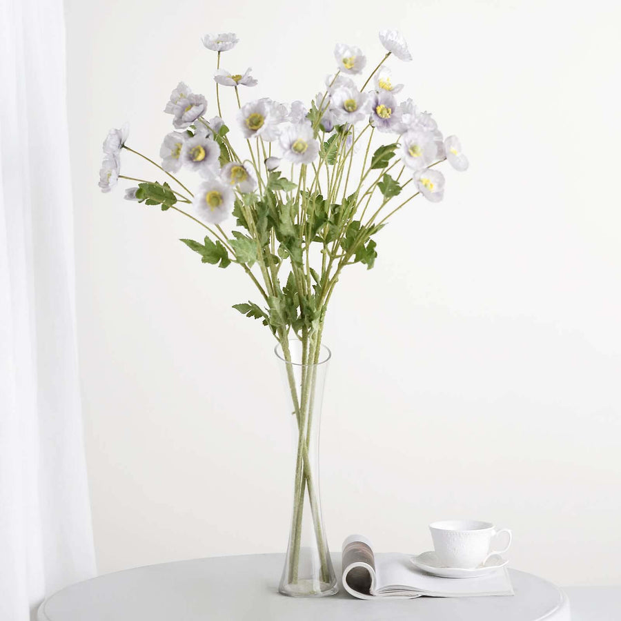 White flower bouquet in a tall vase on a table with a cup and magazine.