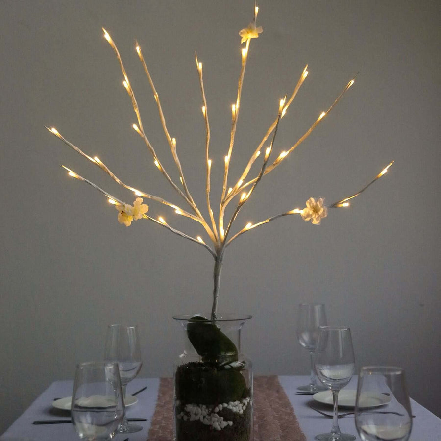 Decorative lighted branch centerpiece with flowers on a dining table.