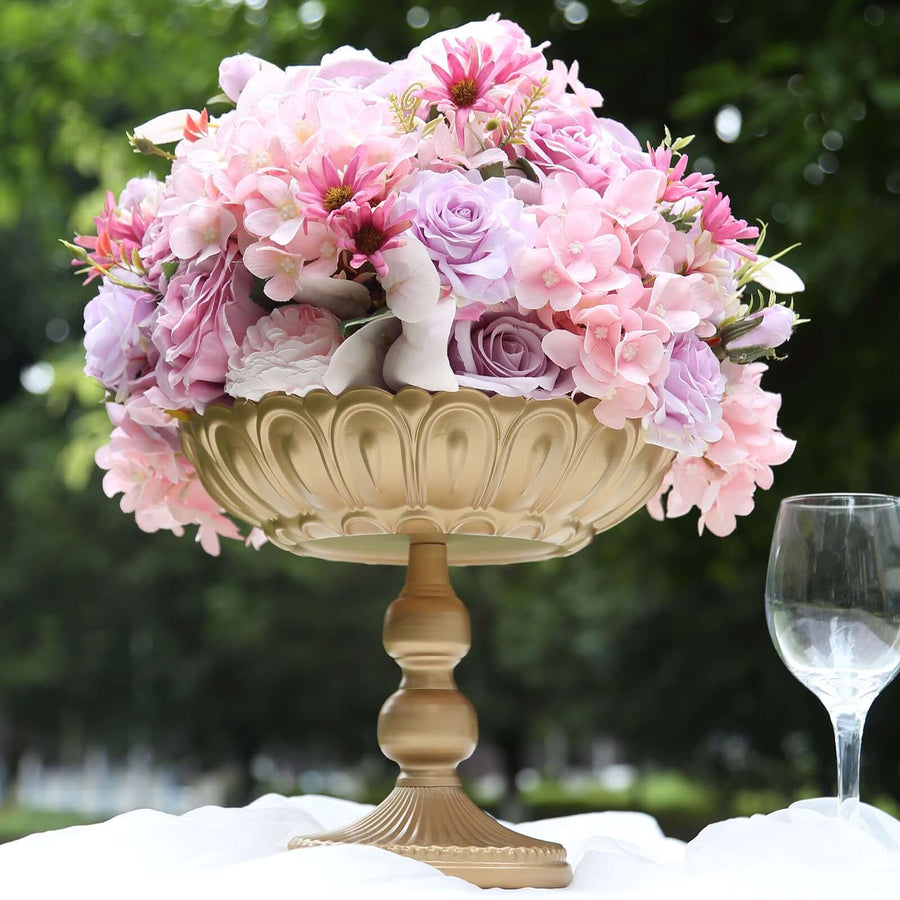 Gold pedestal vase filled with pink and purple flowers, outdoors in a garden setting.