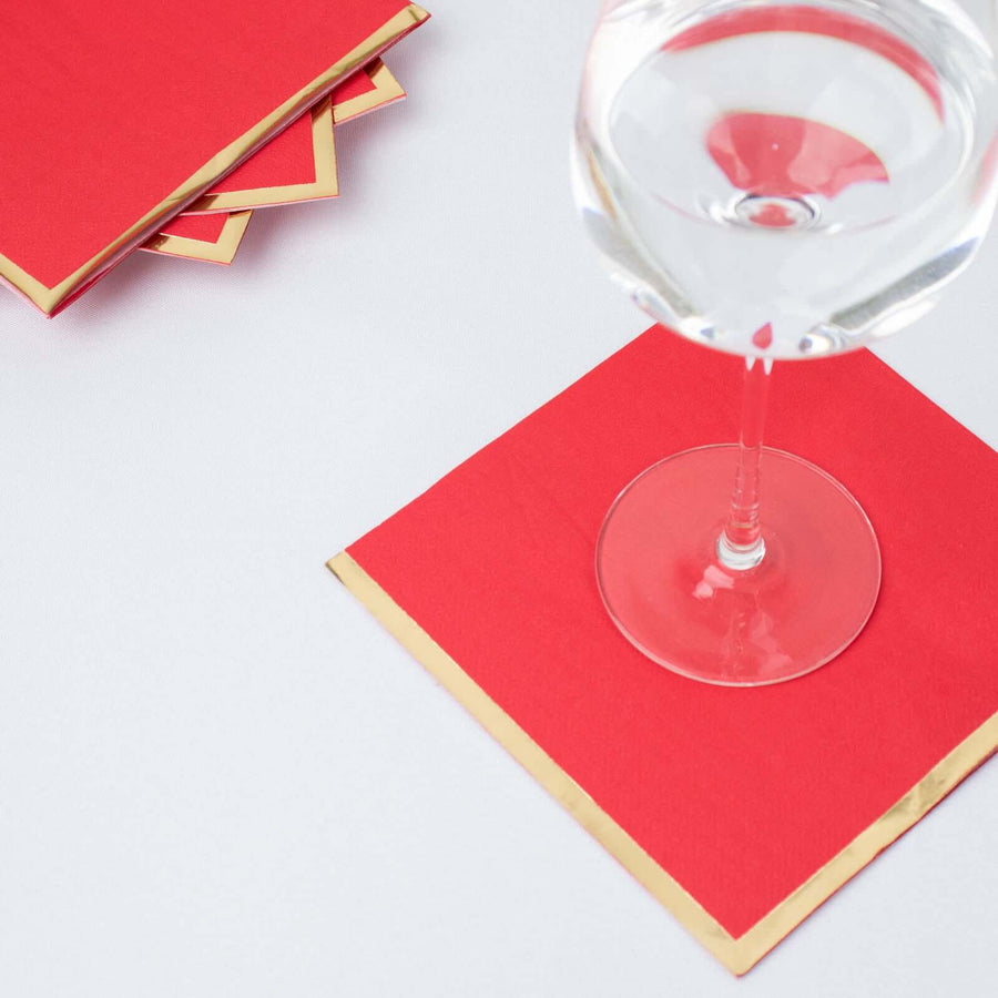Red cocktail napkins with gold trim and a glass on a white surface.