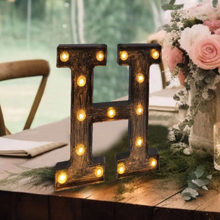 Decorative light-up letter 'H' on a table with flowers in the background.