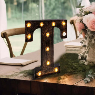 Decorative marquee letter "T" with lights on a wooden table in a floral setting.