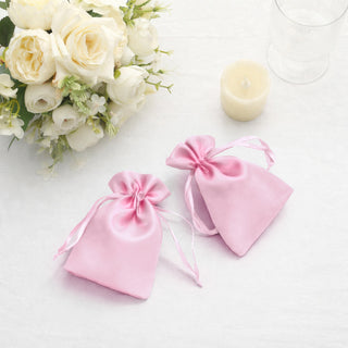 Pink satin drawstring bags on a white table with flowers and candle.