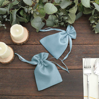 Two blue satin drawstring bags on a rustic wooden table with greenery and candles.