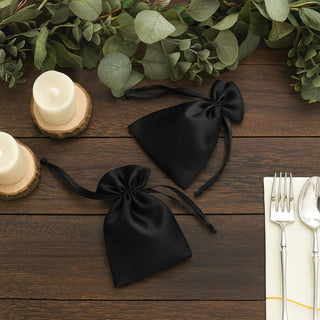 Black satin drawstring bags on a rustic wooden table with candles and greenery.