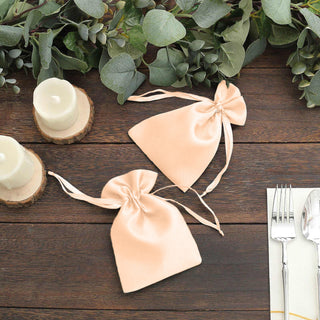 Two peach satin drawstring bags on a wooden table with greenery and candles.