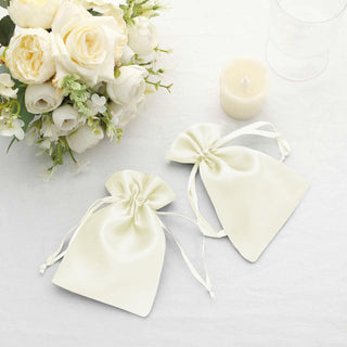 Elegant white satin favor bags beside a floral arrangement and candle.