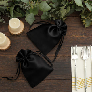 Black satin drawstring bags on a rustic wooden table with candles and greenery.