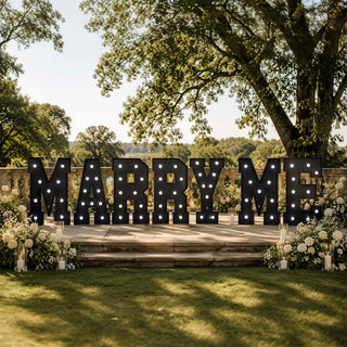 Outdoor wedding marquee sign spelling “Marry Me” with floral decor and candles