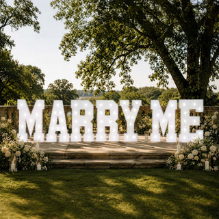 Elegant outdoor wedding sign reading “MARRY ME” with flowers and lights