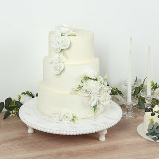 Three-tier wedding cake adorned with white roses, displayed on a decorative pedestal.
