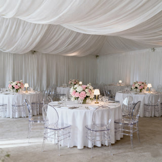 Elegant event setup with floral centerpieces and clear chairs in a decorated tent.