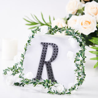 Decorative letter "R" surrounded by greenery and flowers on a white backdrop.