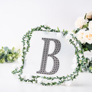 Decorative letter "B" surrounded by greenery and flowers on a white background.