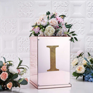 Decorative mirrored box with floral arrangement and gold letter "I" on a textured background.