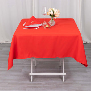 Red tablecloth on a dining table with a glass and vase of flowers.