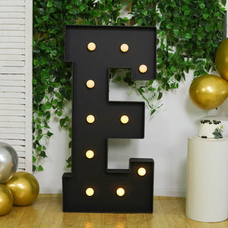 Large decorative letter "E" with lights, set against greenery and balloons.
