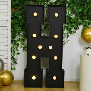 Black marquee letter "H" with lights surrounded by greenery and decor elements.