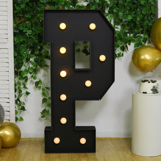 Large illuminated letter "P" with bulbs, set against greenery and festive decor.