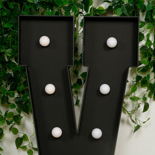 Black marquee letter "V" with white bulbs against a green foliage backdrop.