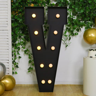 Black marquee letter "V" with glowing bulbs, surrounded by greenery and balloons.
