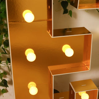 Gold marquee letter with warm light bulbs, surrounded by greenery.