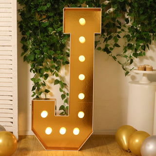 Gold marquee letter "J" illuminated with bulbs, surrounded by greenery and balloons.
