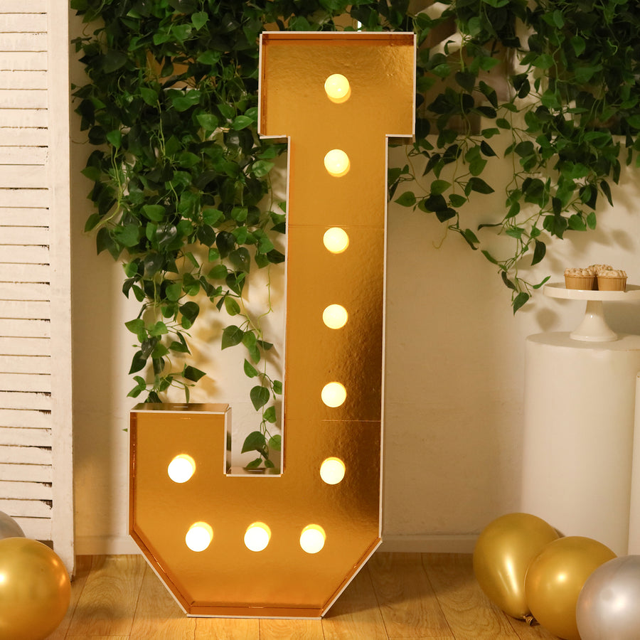 Gold marquee letter "J" illuminated with bulbs, surrounded by greenery and balloons.