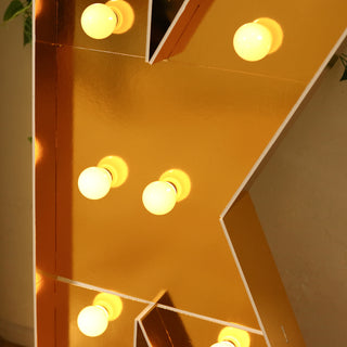 Gold marquee letter with glowing bulbs on a plain background.