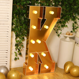 Gold marquee letter "K" with lightbulbs, surrounded by greenery and balloons.