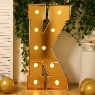 Gold marquee letter "K" with lights, surrounded by greenery and balloons.