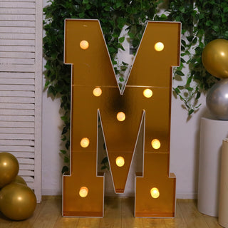 Gold marquee letter "M" with lights, surrounded by greenery and balloons.