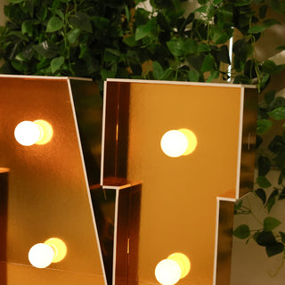 Gold marquee letter with lights reflecting, surrounded by greenery.
