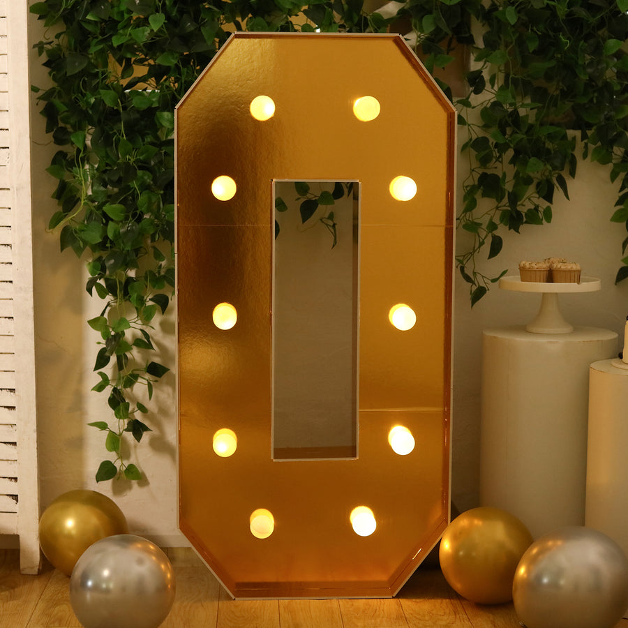 Gold marquee number zero with lights, surrounded by greenery and balloons.