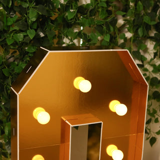 Gold marquee letter with lights against a green leafy backdrop.