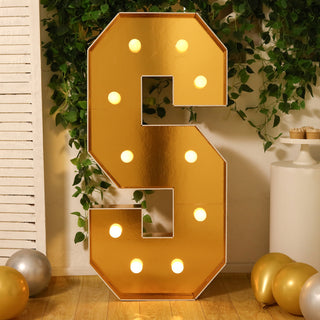 Gold marquee letter "S" with lights, surrounded by greenery and balloons.