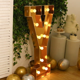 Gold marquee letter "Y" with lights against greenery backdrop.
