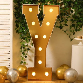 Gold marquee letter "Y" with lights, surrounded by greenery and balloons.