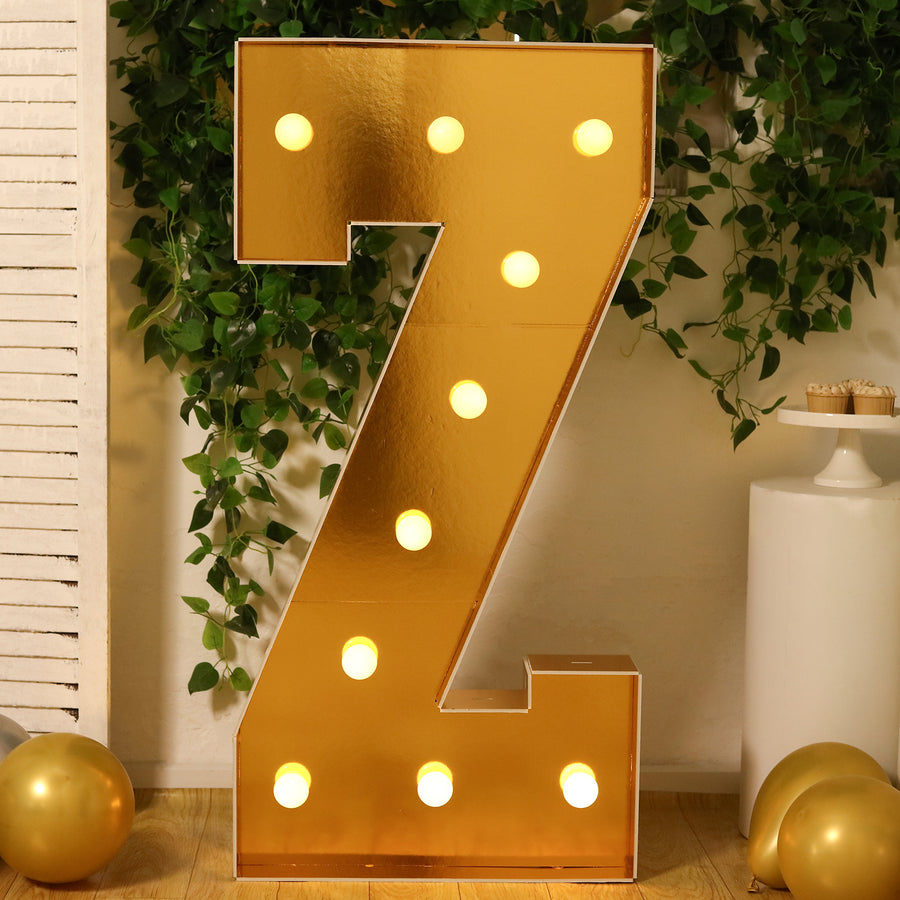 Gold marquee letter Z with lights, surrounded by greenery and balloons.