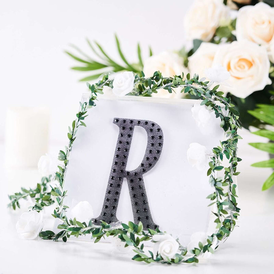 Black letter "R" surrounded by greenery and white flowers on a soft background.