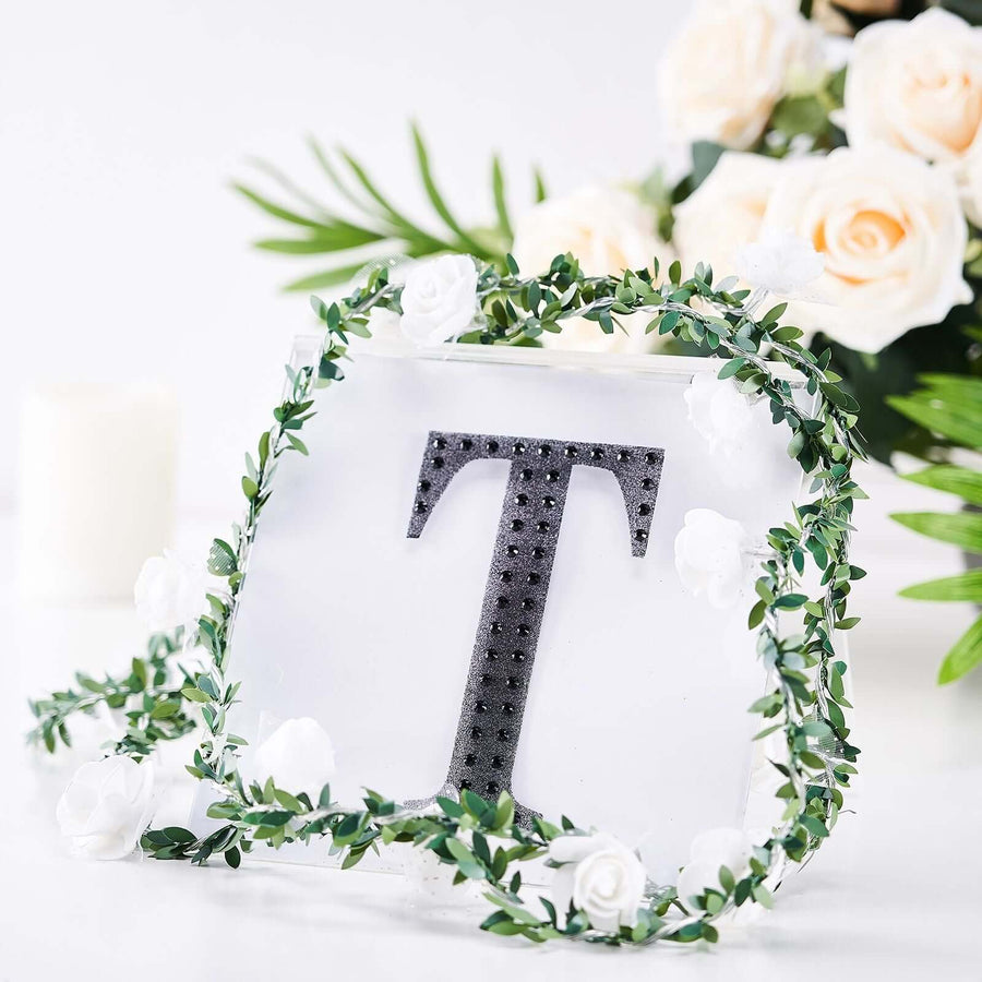 Decorative letter "T" with rhinestones surrounded by greenery and white flowers.