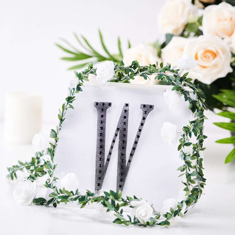 Decorative letter "W" with rhinestones surrounded by greenery and flowers.