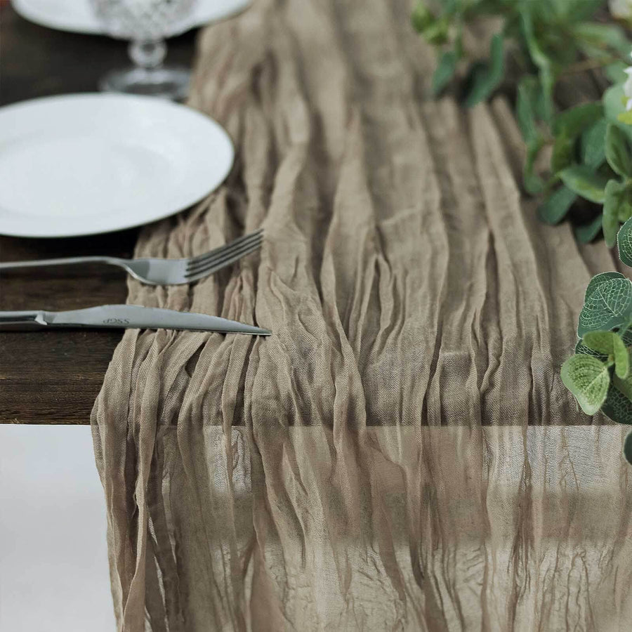 Wrinkled beige table runner on a wooden table with plates and greenery.