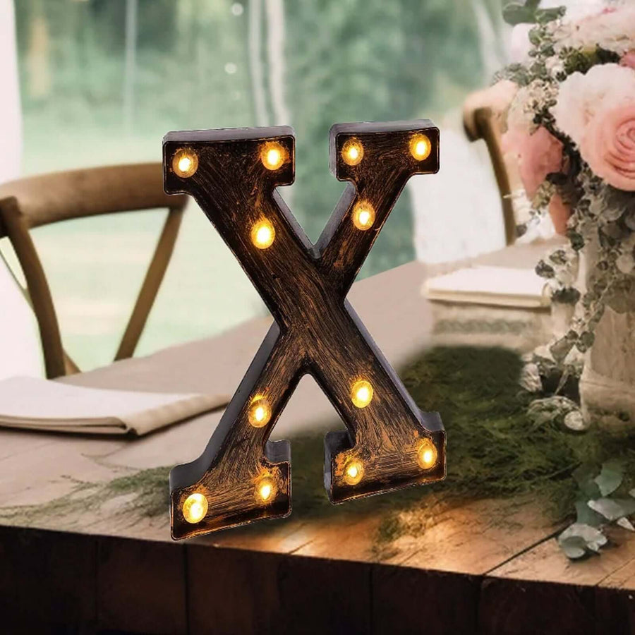 Decorative light-up letter "X" on a rustic table with flowers and greenery.