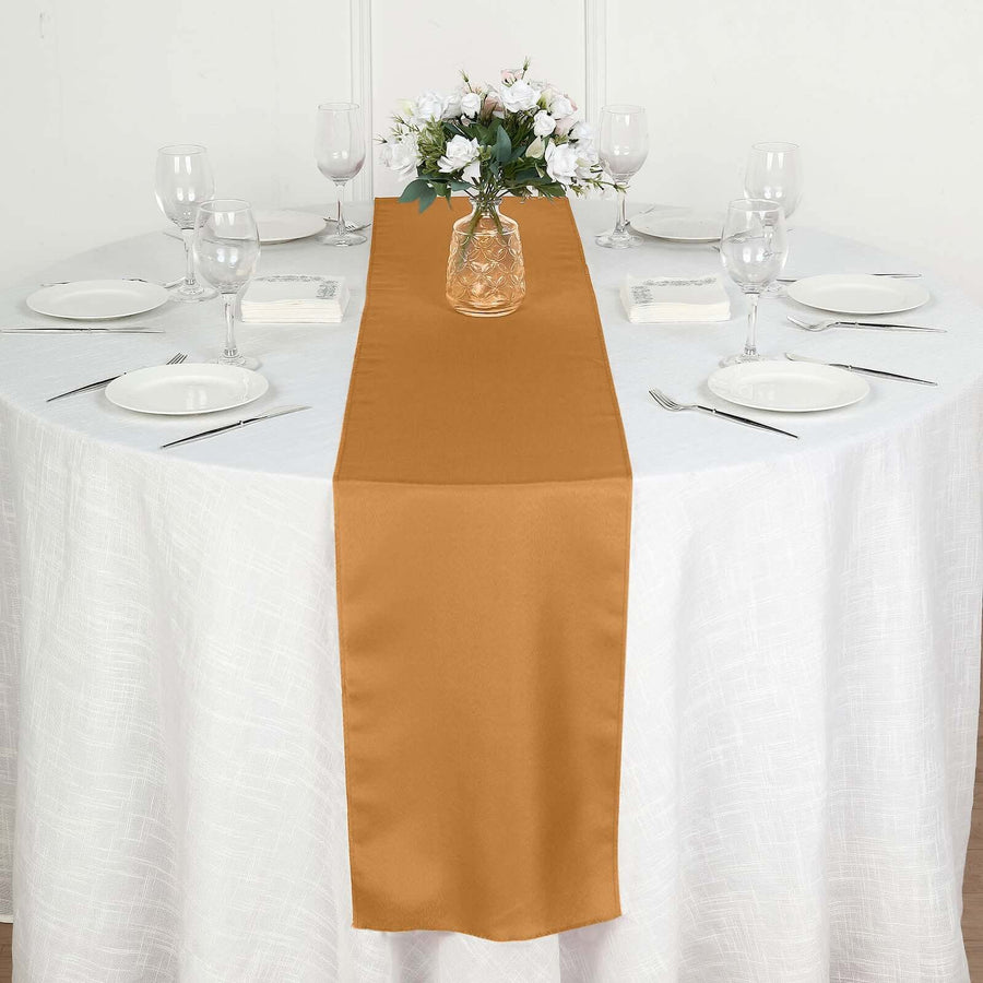 Gold satin table runner on a white table with glassware and floral arrangement.