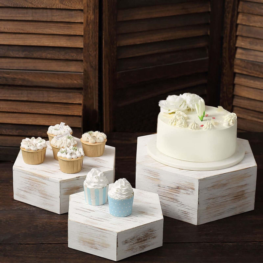 Elegant white cake with floral decoration and assorted cupcakes on rustic hexagonal stands.