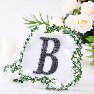 Black letter "B" surrounded by greenery and flowers on a white background.